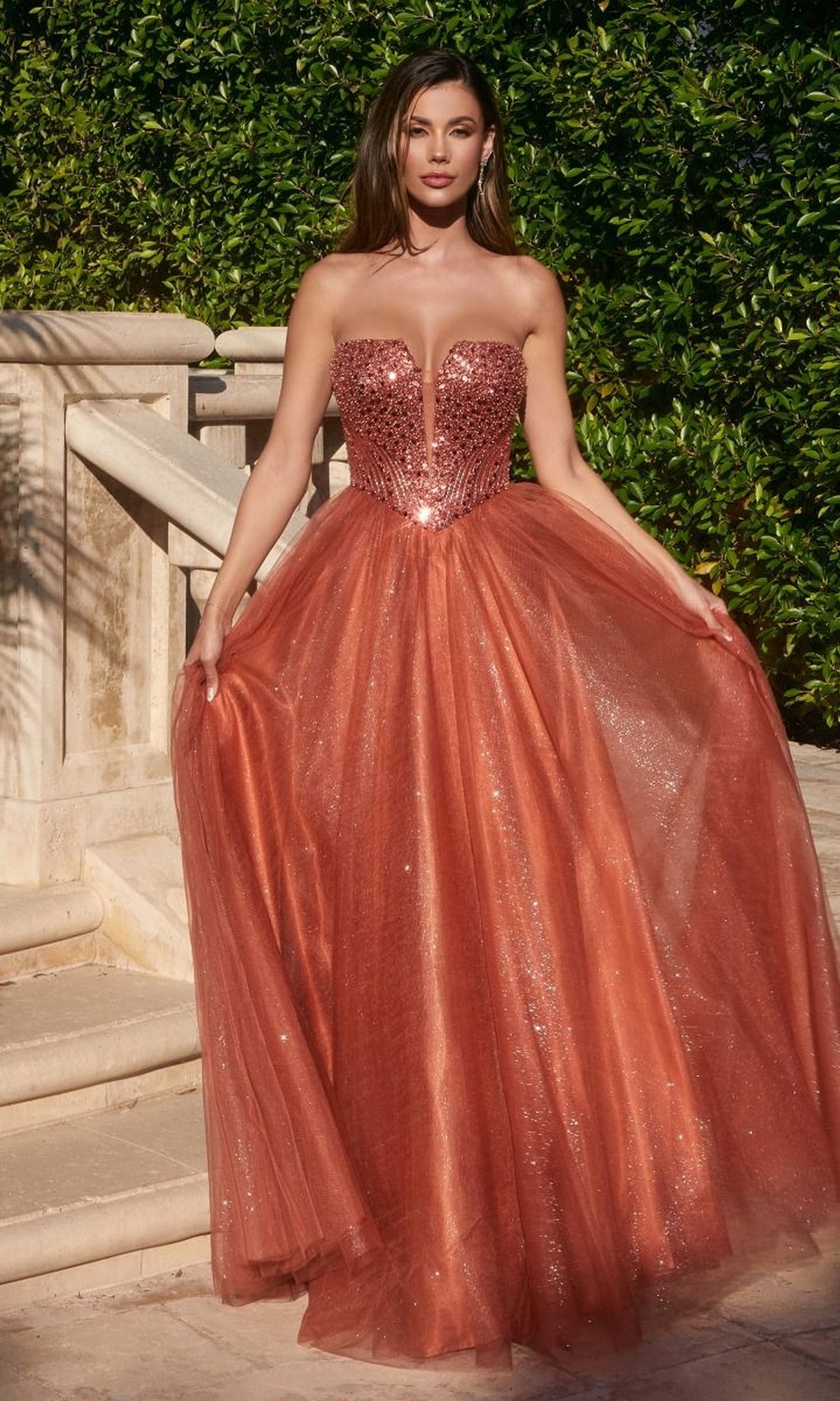 A woman stands outdoors on stone steps, wearing the Lace-Up Strapless Long Glitter Prom Dress CD0217—a floor-length, shimmery copper gown with a fitted bodice and full tulle skirt.